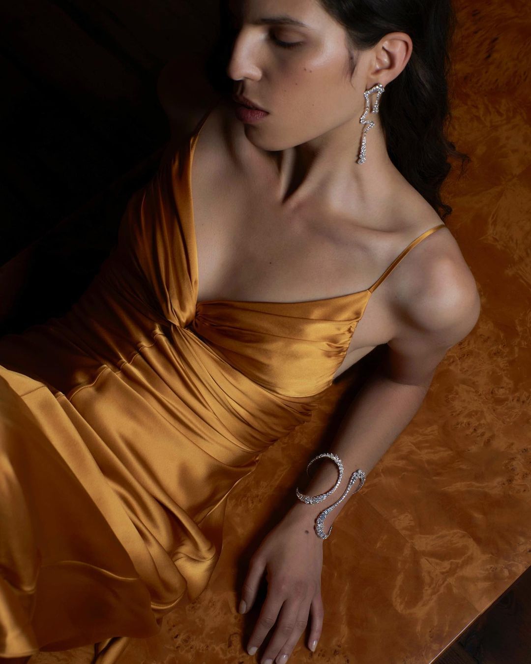 Woman poses wearing a golden dress with jewelry, including earrings and a bracelet, in a dark indoor setting with warm lighting.