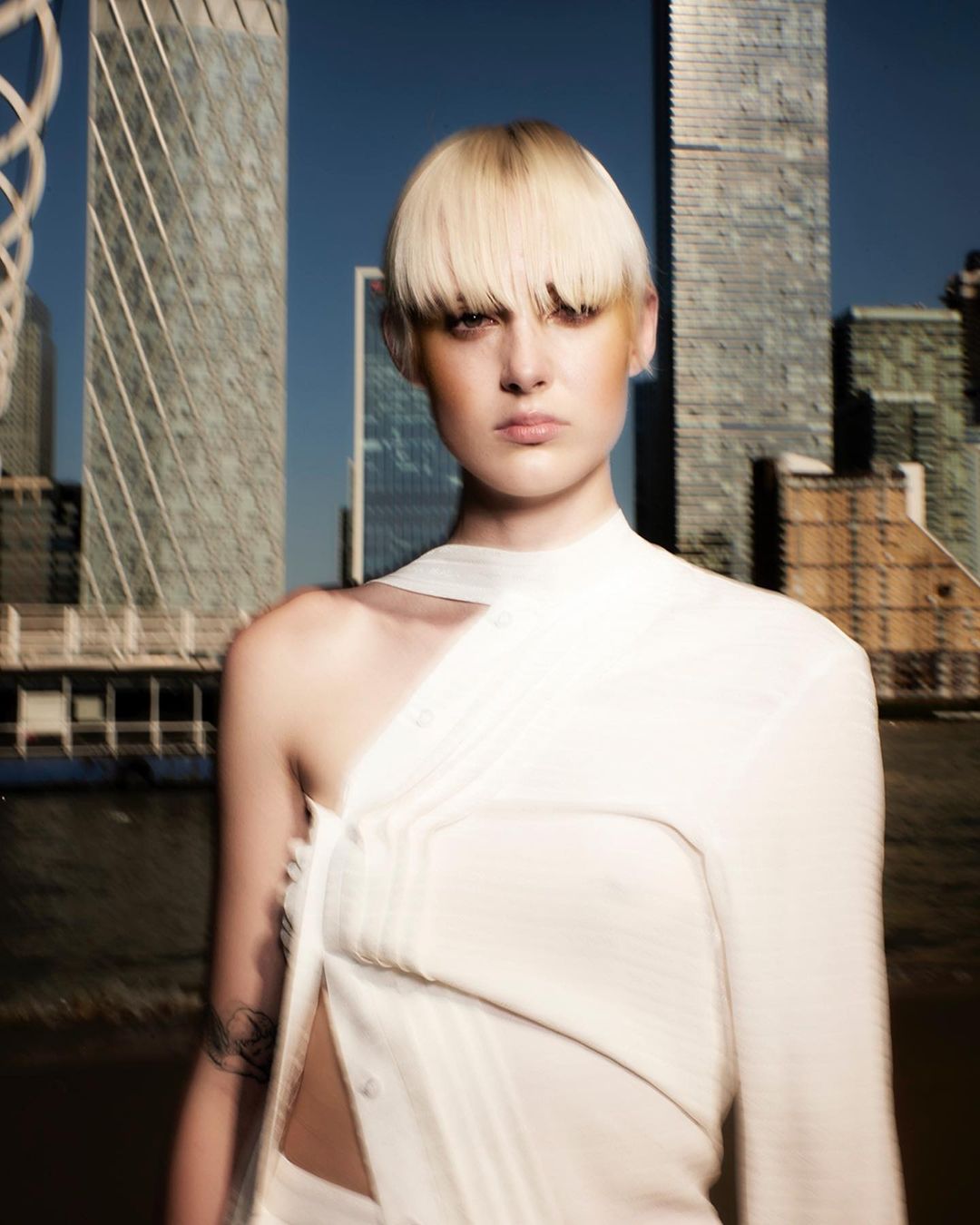 Close-up of a woman with light blonde hair and a modern top posing in front of an urban cityscape on a clear day.
