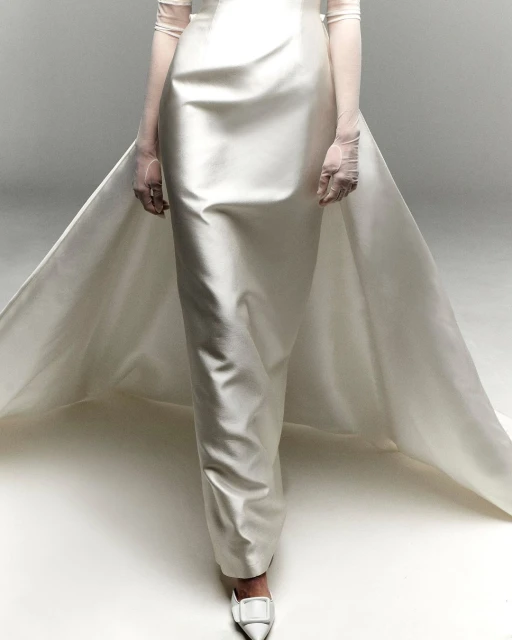 A person wears a white satin gown with a train and matching heels while posing in a photo studio with a neutral white background.