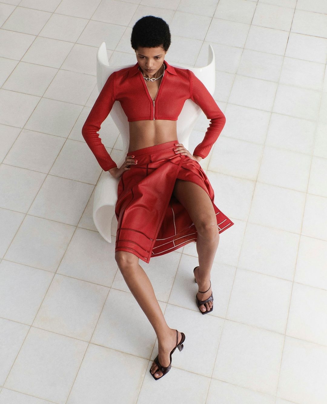 Model wearing a matching red zip-up crop top and red skirt seated in a white chair against a tiled floor.