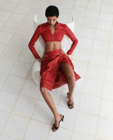 Model wearing a matching red zip-up crop top and red skirt seated in a white chair against a tiled floor.