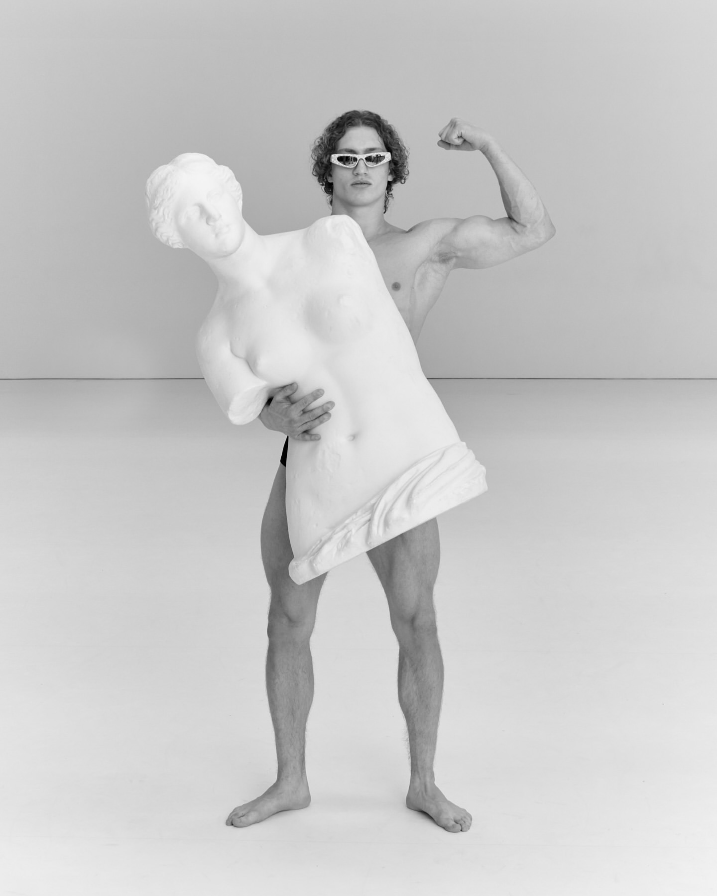 A muscular man in goggles strikes a pose, holding a broken classical statue of a woman in an indoor studio with a gray background.