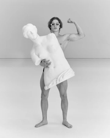A muscular man in goggles strikes a pose, holding a broken classical statue of a woman in an indoor studio with a gray background.