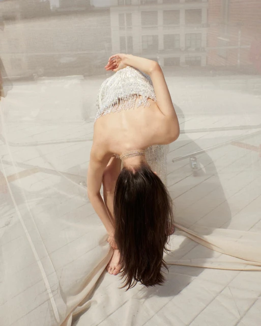 Brunette woman in a fringed dress bends backward on a rooftop, showcasing a blend of fashion and architectural setting in soft lighting.
