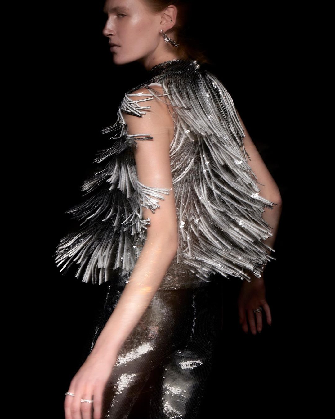 Woman in shimmering silver top with fringe detail and metallic trousers against a dark backdrop, fashion editorial photography.
