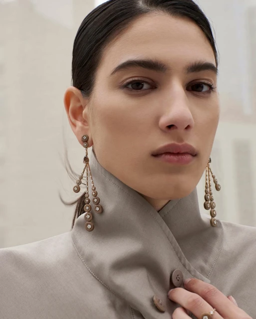 Portrait of an adult woman wearing chandelier earrings and a neutral-colored jacket with a ring on her finger against a neutral background.