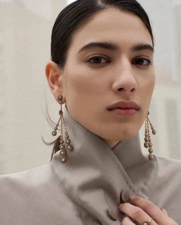 Portrait of an adult woman wearing chandelier earrings and a neutral-colored jacket with a ring on her finger against a neutral background.