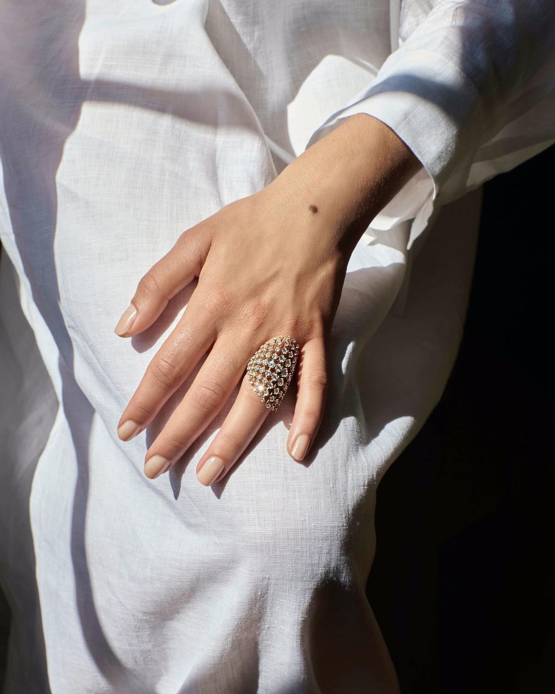 Close-up of a female hand with a large gold geometric ring, wearing a white linen shirt with soft natural light and shadows.