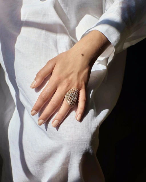 Close-up of a female hand with a large gold geometric ring, wearing a white linen shirt with soft natural light and shadows.