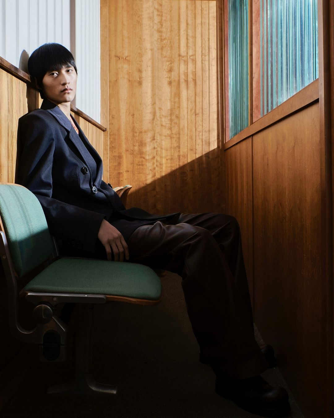 Young adult in dark suit sits on a green seat inside wood-paneled room with colorful window panes, looking seriously at the camera.