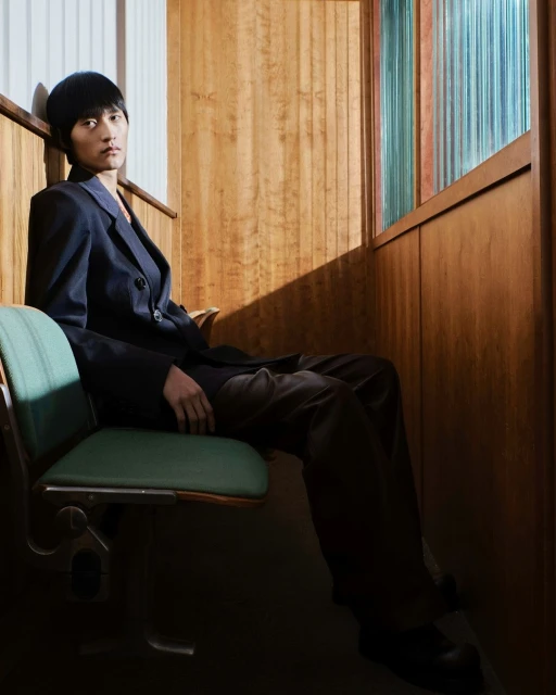 Young adult in dark suit sits on a green seat inside wood-paneled room with colorful window panes, looking seriously at the camera.