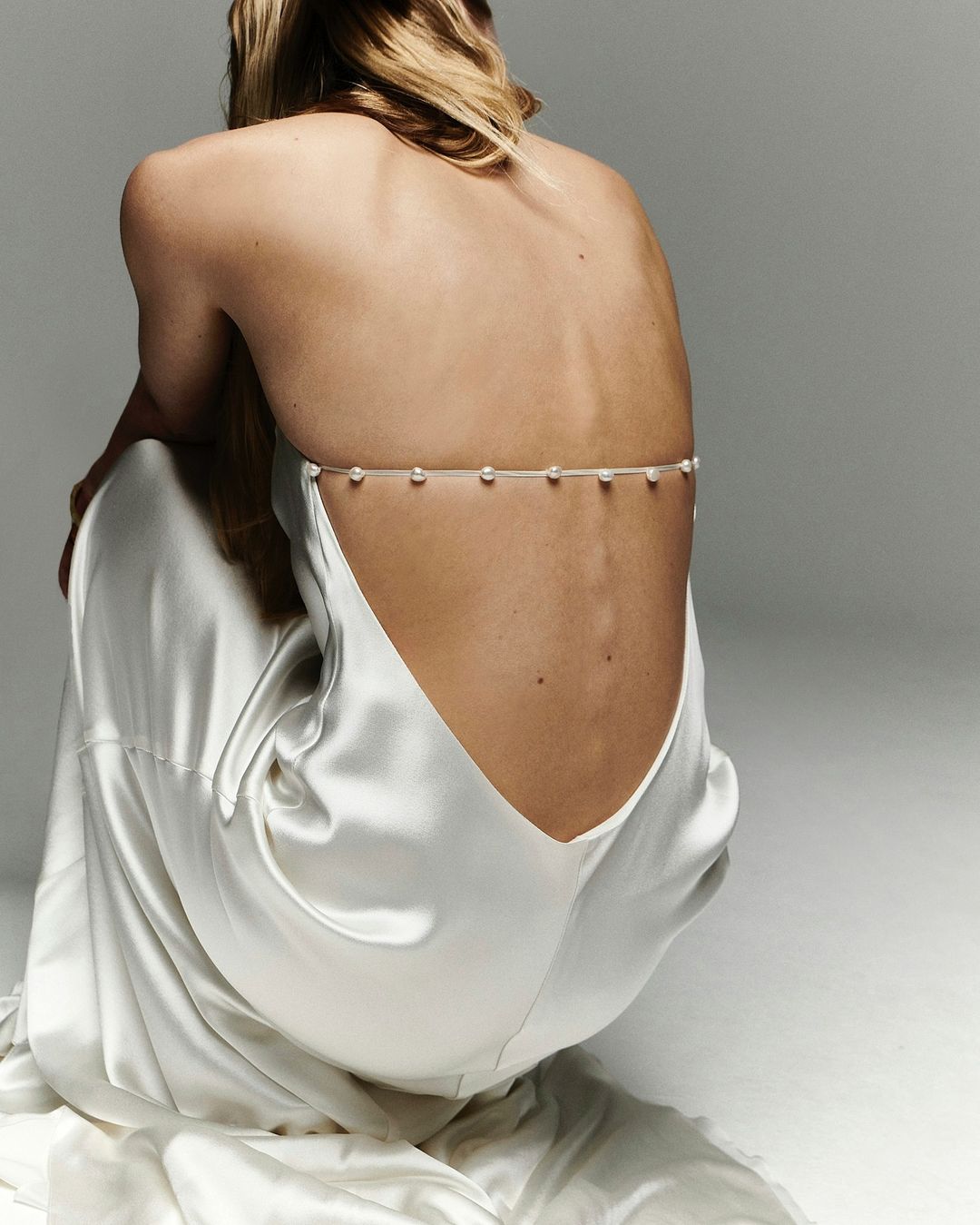 Back view of a woman with blonde hair wearing a backless white satin dress and pearl body jewelry, posing against a grey backdrop.