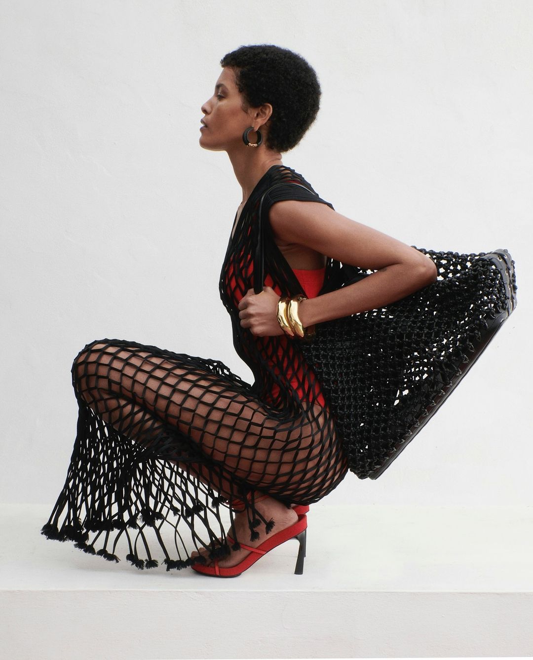 Elegant woman with afro in black fishnet dress and red heels poses in a photo studio with a black woven bag and gold bracelets.