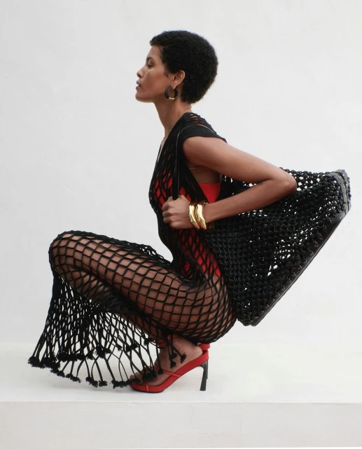 Elegant woman with afro in black fishnet dress and red heels poses in a photo studio with a black woven bag and gold bracelets.