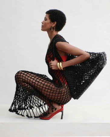 Elegant woman with afro in black fishnet dress and red heels poses in a photo studio with a black woven bag and gold bracelets.