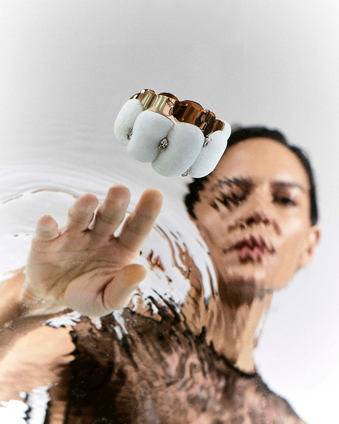 Close-up portrait of a woman reaching toward the camera through a rippled water surface with a metallic brass knuckle ring visible above.