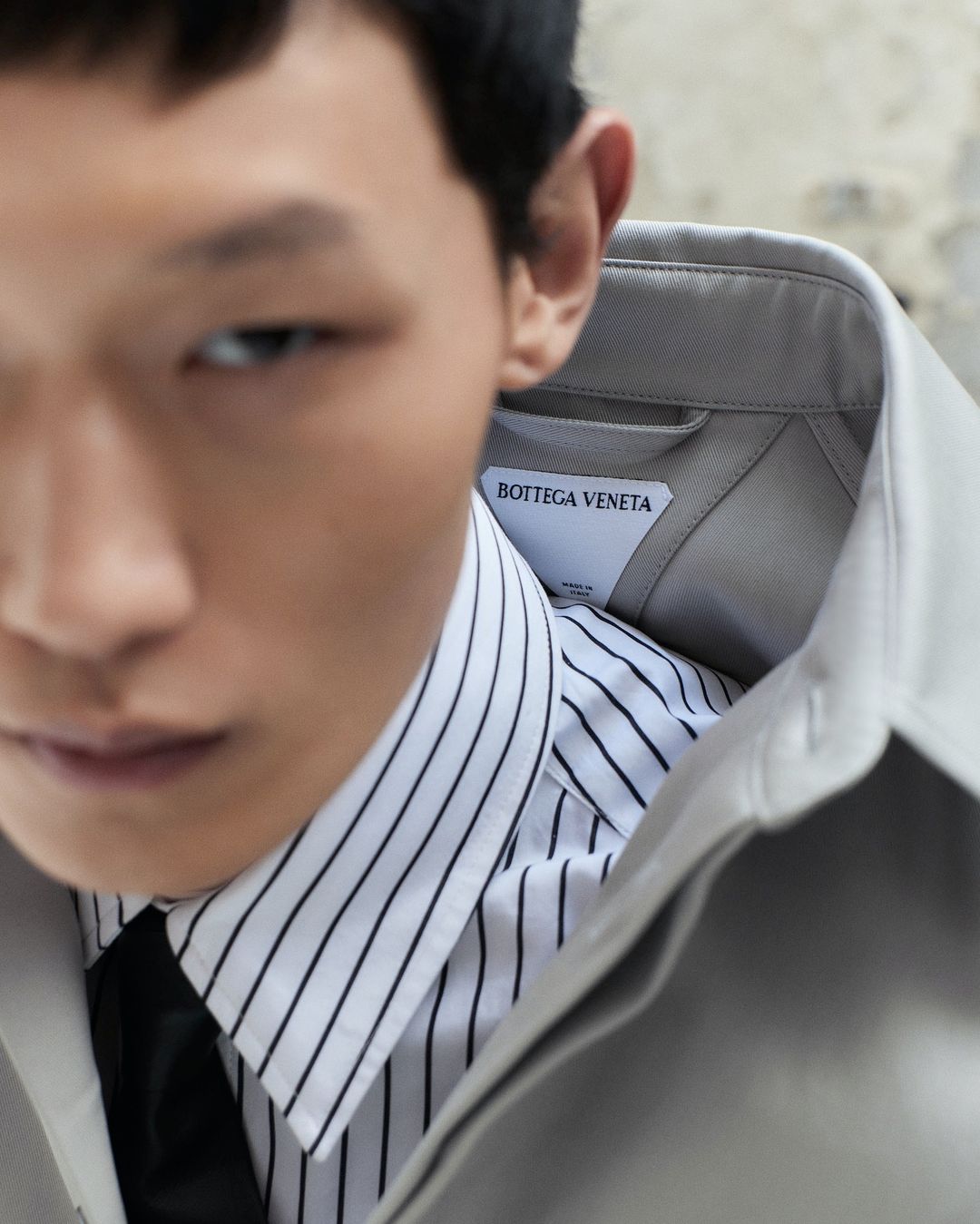 Close-up portrait of a male model in a striped shirt, black tie, and tan jacket, featuring a Bottega Veneta label.