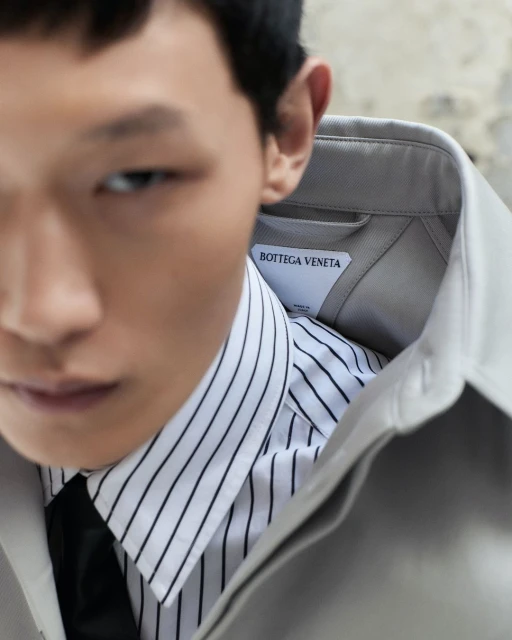 Close-up portrait of a male model in a striped shirt, black tie, and tan jacket, featuring a Bottega Veneta label.