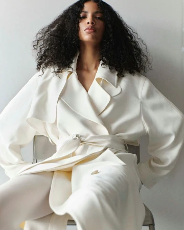 Model with long dark curly hair wearing a white belted coat seated on a chair against a neutral wall.