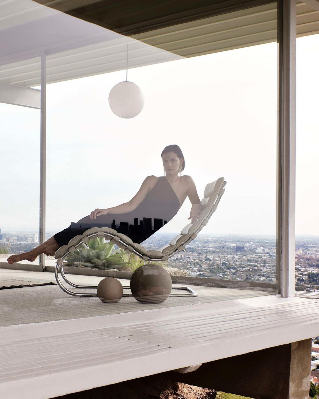 Woman in gray dress lounges on modern chaise lounge with ball-shaped objects indoors, cityscape and sky visible through windows.