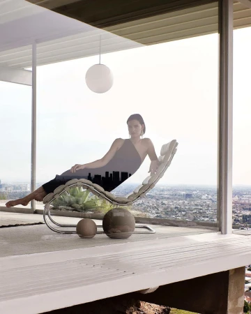 Woman lounging on a chrome chair inside a modern glass-walled room with a city skyline silhouette superimposed on her dress.