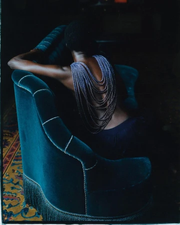 Rear view of a person with short dark hair wearing layered beaded necklaces draped across their bare back while leaning against a dark teal velvet armchair in London by Theresa Marx