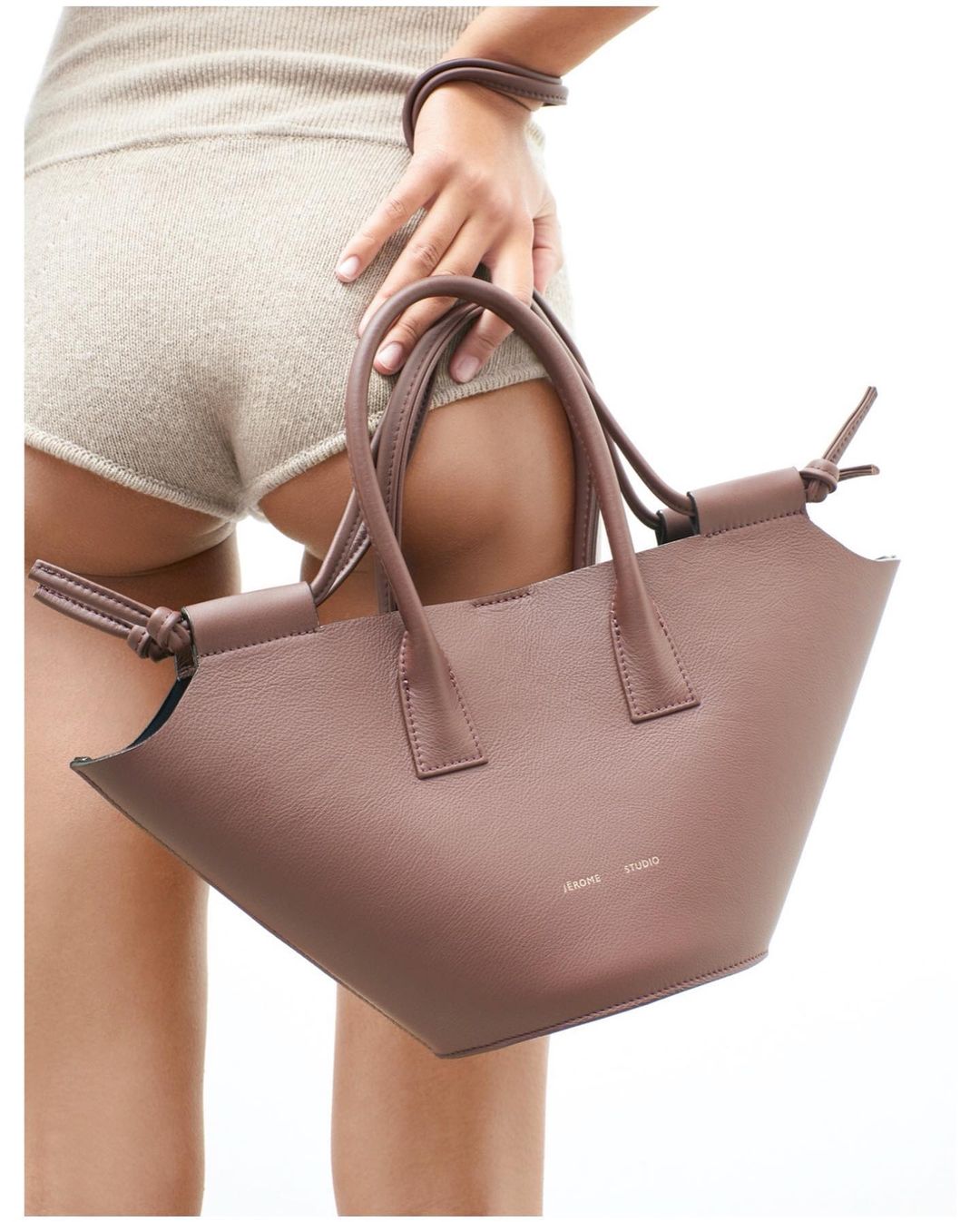Close-up of a tan leather handbag with a tote style and short handles, being held by an adult female wearing casual beige clothing.