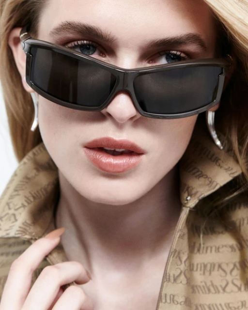Close-up shot of a blonde woman wearing sunglasses and a patterned shirt. The photo studio lighting emphasizes her features.