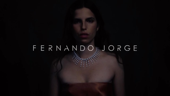 Elegant woman with fair skin and shoulder-length dark hair, wearing a strapless dress and pearl necklace, lit against a dark background.