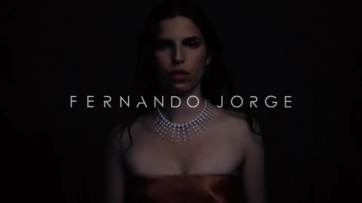 Elegant woman with fair skin and shoulder-length dark hair, wearing a strapless dress and pearl necklace, lit against a dark background.