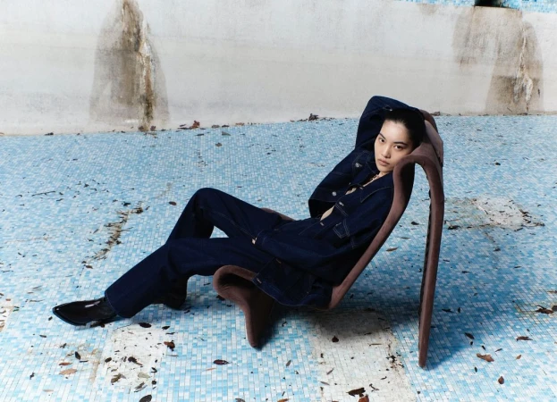Fashion shot of a young Asian woman in a denim jacket and jeans reclining in an armless chair on a cracked blue tile surface.