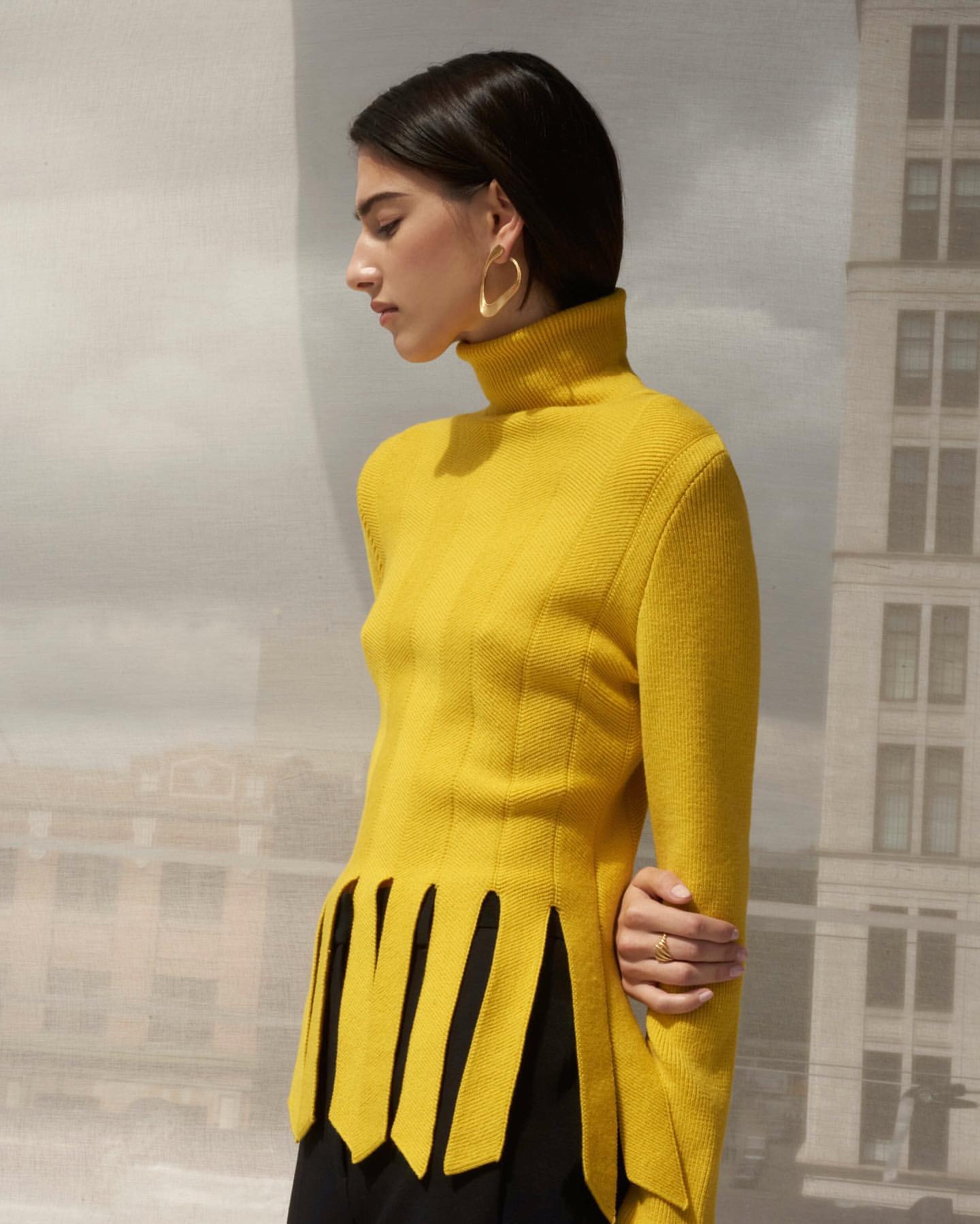 Woman in yellow turtleneck sweater poses against backdrop of building. Fashion shot, eye-level angle, indoor setting.