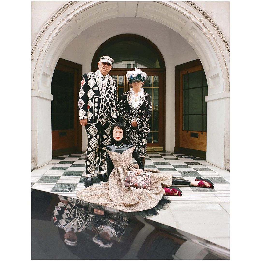 Three people stand outside a building; two wear pearl-covered suits and hats, and one sits in front in a patterned dress and clutch.