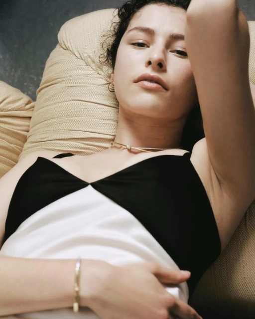 Portrait of a young adult woman lying on a light brown couch wearing a black and white tank top, a bracelet and a necklace.