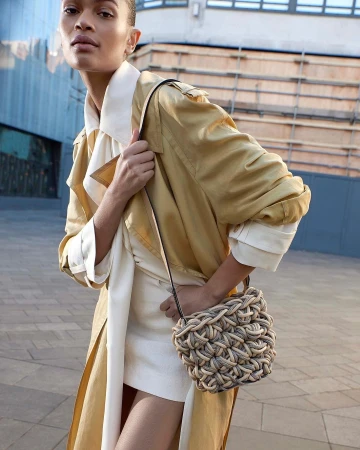 Model wearing a yellow trench coat and white dress carrying a woven rope handbag outdoors