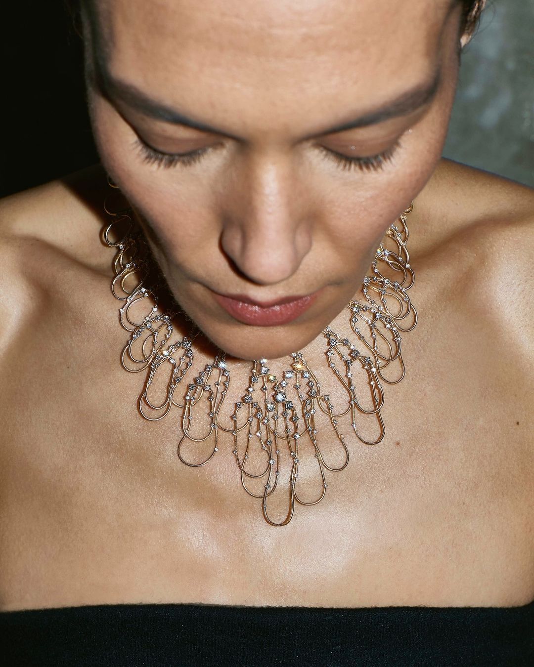 Close-up studio shot of an adult woman wearing a metallic necklace with clear gemstones, focusing on her face and jewelry.