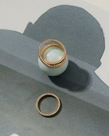 Elevated jewelry product shot showcases gold diamond rings, one encircling a pale blue sphere, on a muted gray, white, and blue patterned surface.