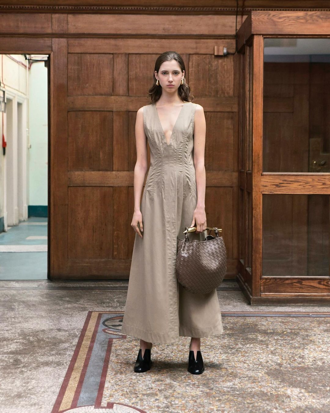 A woman in a dress stands in an interior with wood panels holding a brown woven bag. She is wearing black heeled shoes.