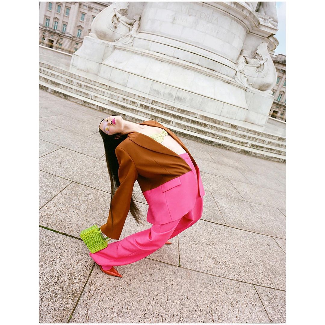 Woman in brown blazer and pink pants poses with a green handbag in front of a marble monument and steps, fashion editorial style.