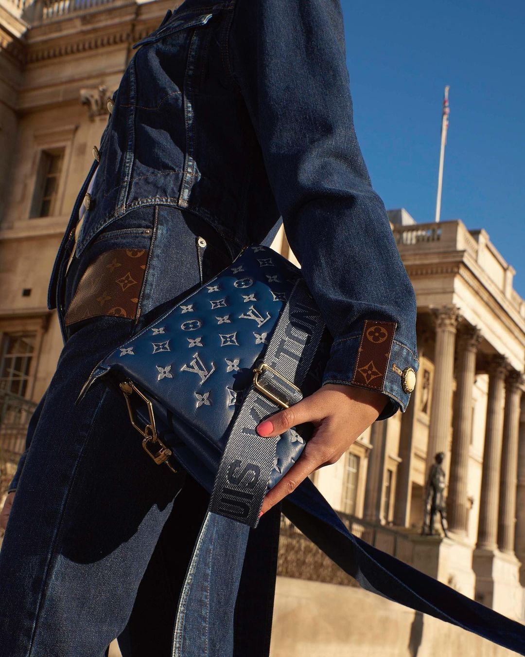 Person in denim with Louis Vuitton bag in front of building with columns under blue sky in fashion or travel editorial shoot.