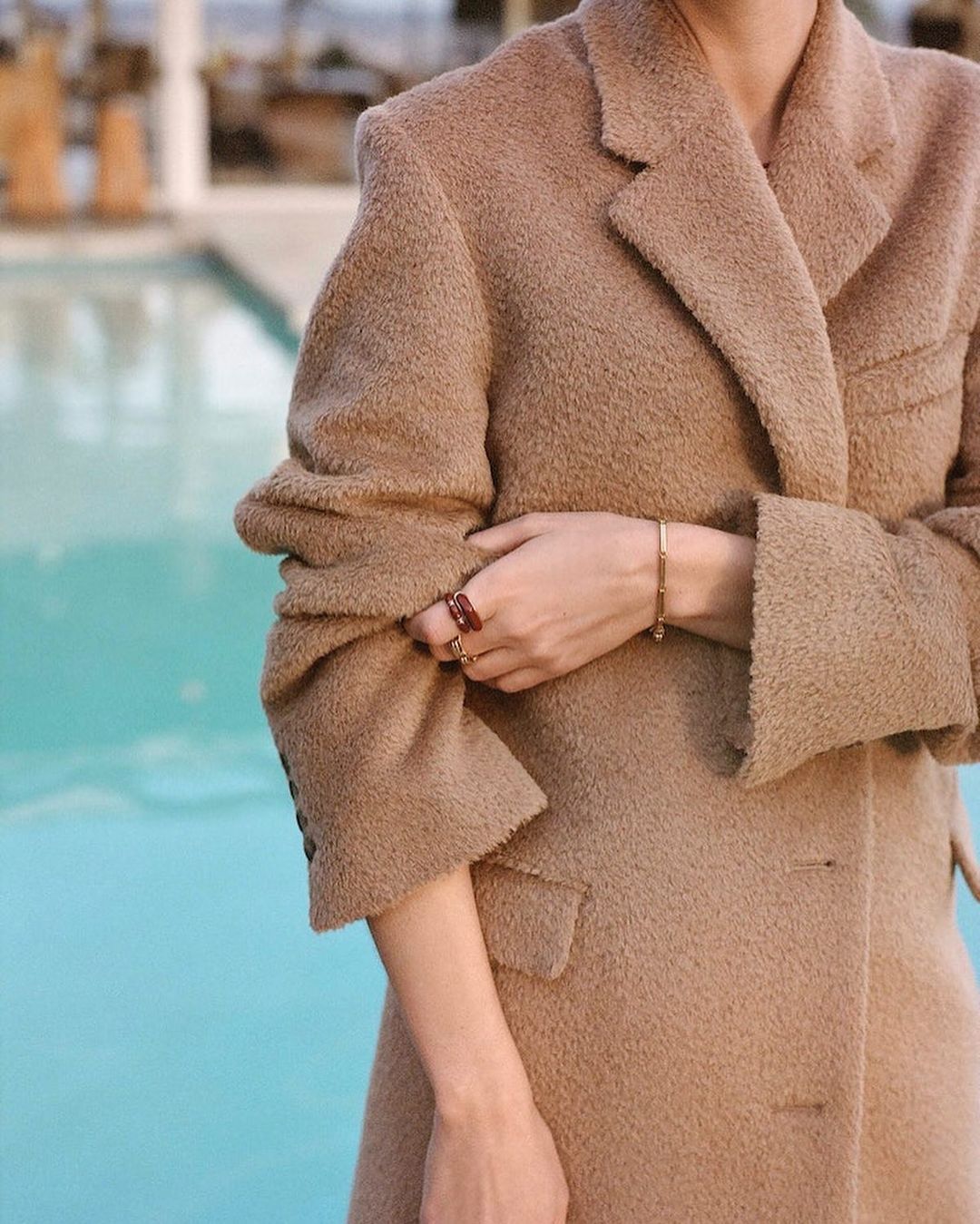 Close-up shot of an adult female in a camel-colored, oversized wool coat, standing against a turquoise pool backdrop with blurred out-of-focus background.
