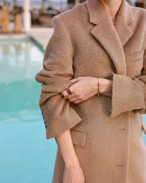 Close-up shot of an adult female in a camel-colored, oversized wool coat, standing against a turquoise pool backdrop with blurred out-of-focus background.