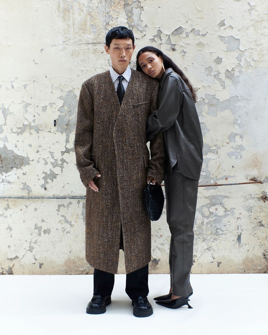 An Asian male and a mixed-race female pose indoors against a distressed wall, modeling in a fashion editorial. The male wears a long overcoat.