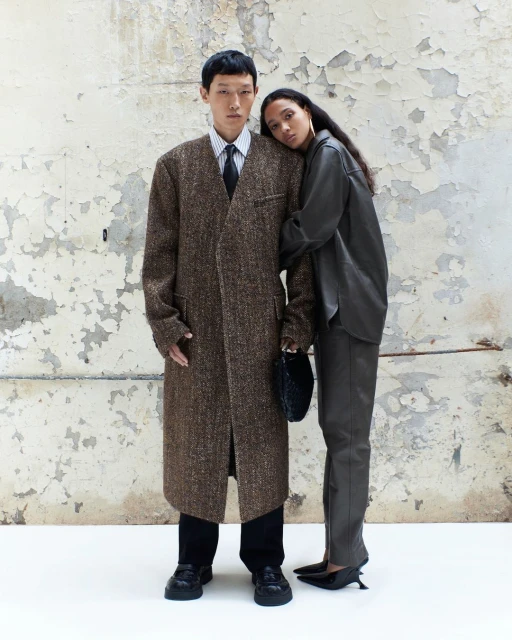 An Asian male and a mixed-race female pose indoors against a distressed wall, modeling in a fashion editorial. The male wears a long overcoat.