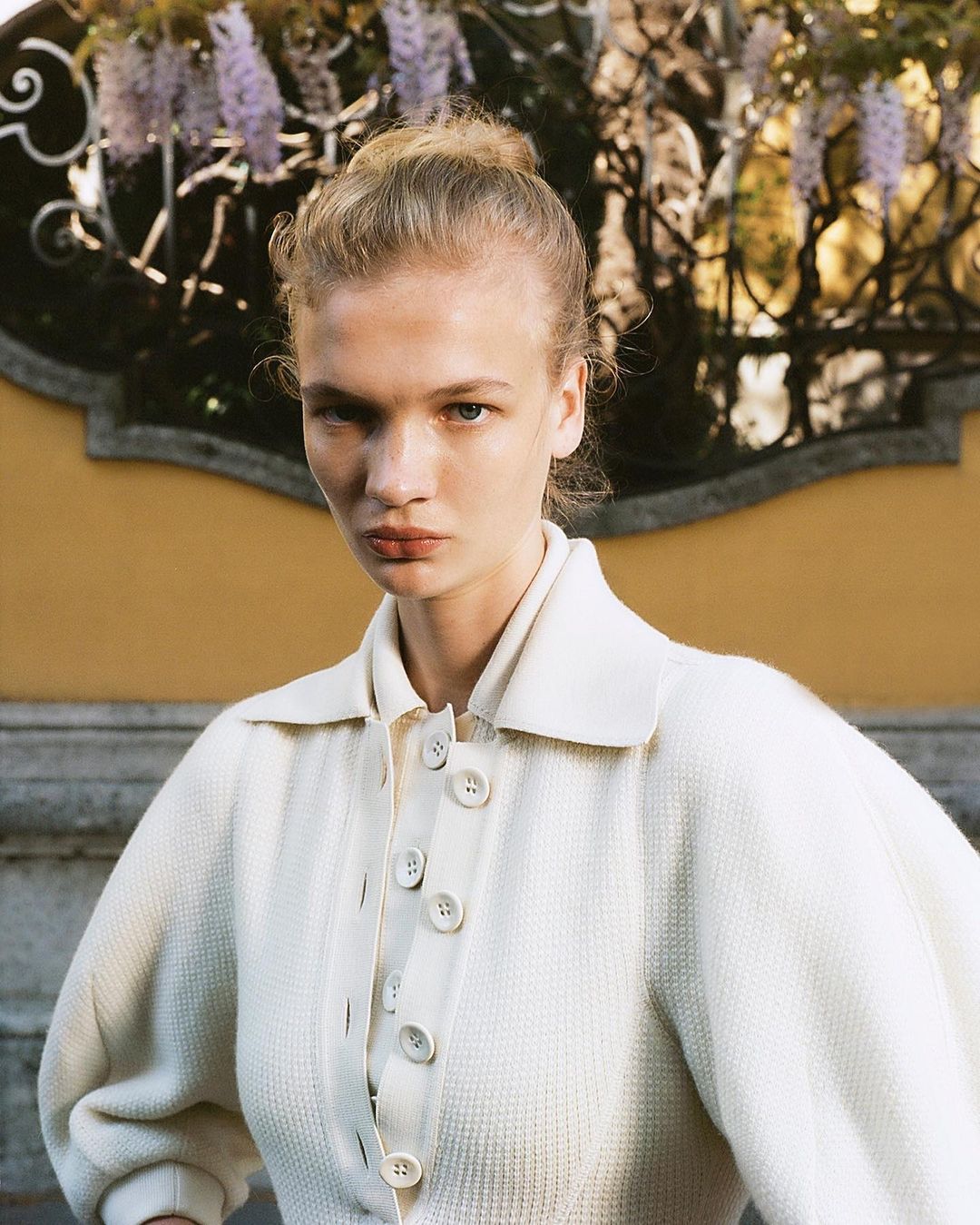 Close-up of a fair-skinned young woman with a neutral expression, wearing a collared white jacket with buttons, in front of flowering vines.