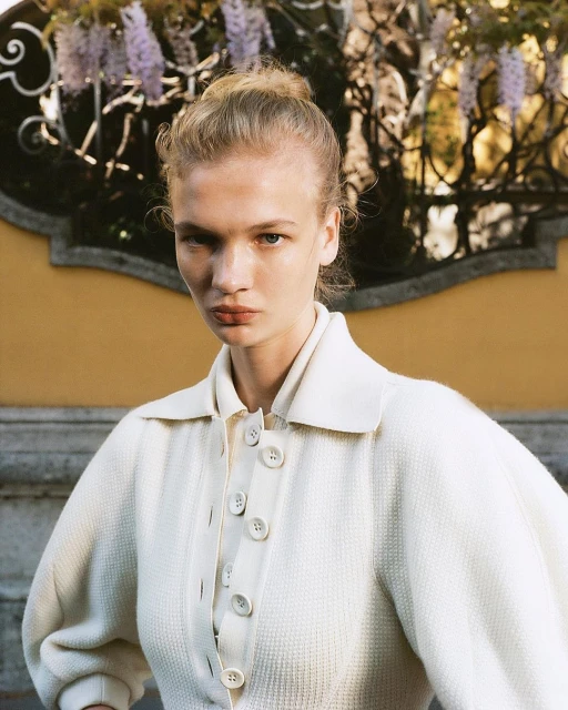 Close-up of a fair-skinned young woman with a neutral expression, wearing a collared white jacket with buttons, in front of flowering vines.