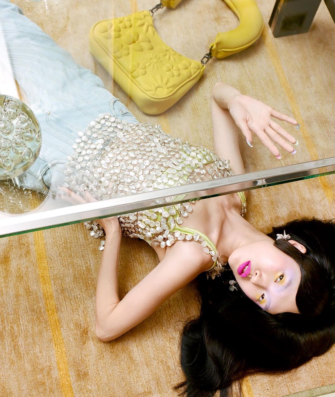 Model lying on patterned carpet wearing sequined top with yellow handbag nearby