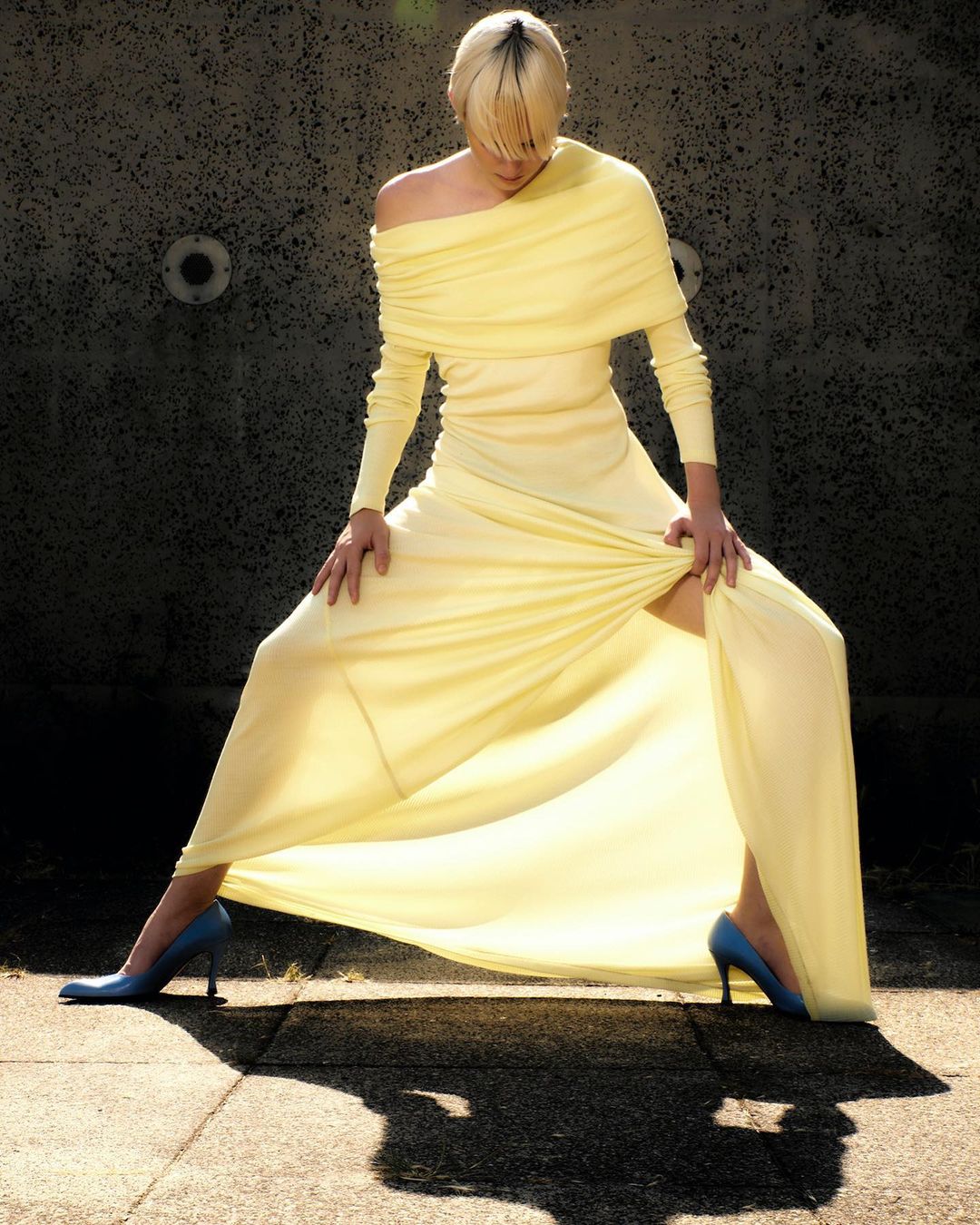 A woman in a long pale yellow dress stands with legs apart on a sidewalk with a grey wall as a backdrop, creating a dynamic pose.