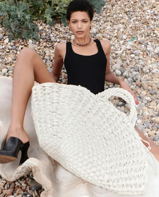 A woman in a black swimsuit poses with a large woven beach bag on a pebble beach, wearing black high heel mules and a gold necklace.