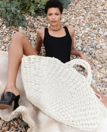 A woman in a black swimsuit poses with a large woven beach bag on a pebble beach, wearing black high heel mules and a gold necklace.